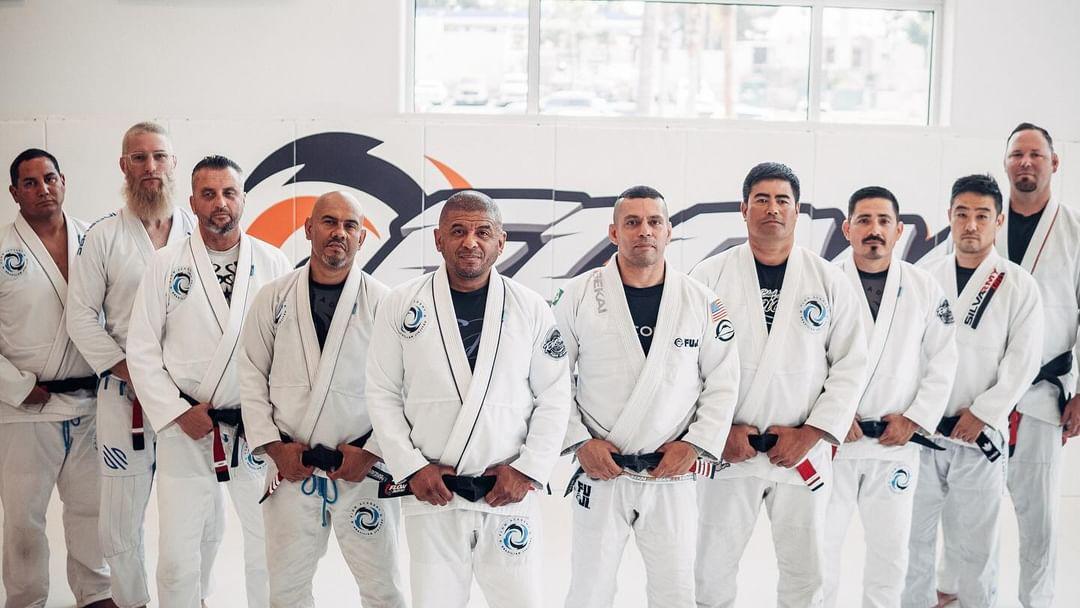A row of black belts in white Flow Academy gis standing in front of the orange Flow wall logo