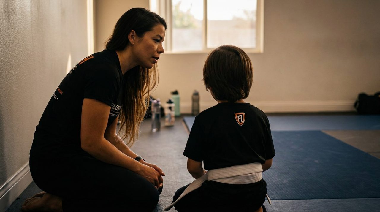 A coach kneeling beside a young student near a quiet corner of the mat before a Flow Limitless class begins, the child seen from behind.