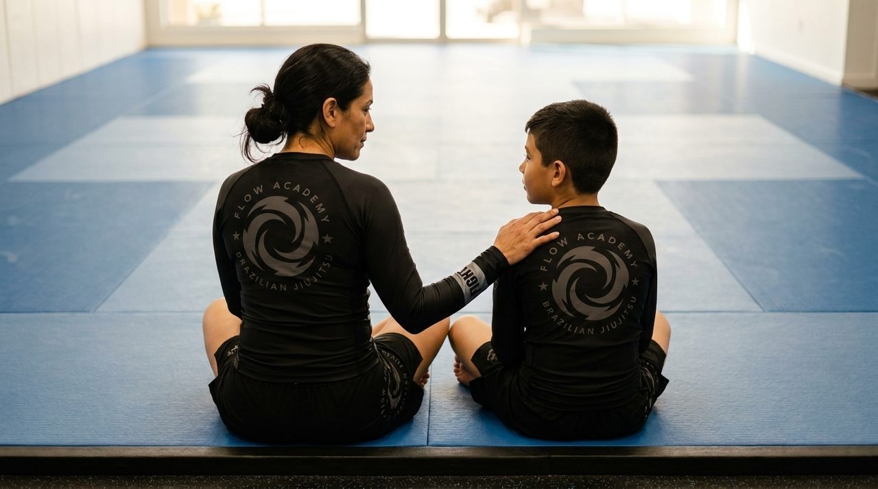 A woman and a boy sit side by side on an empty mat, seen from behind, her hand on his shoulder.