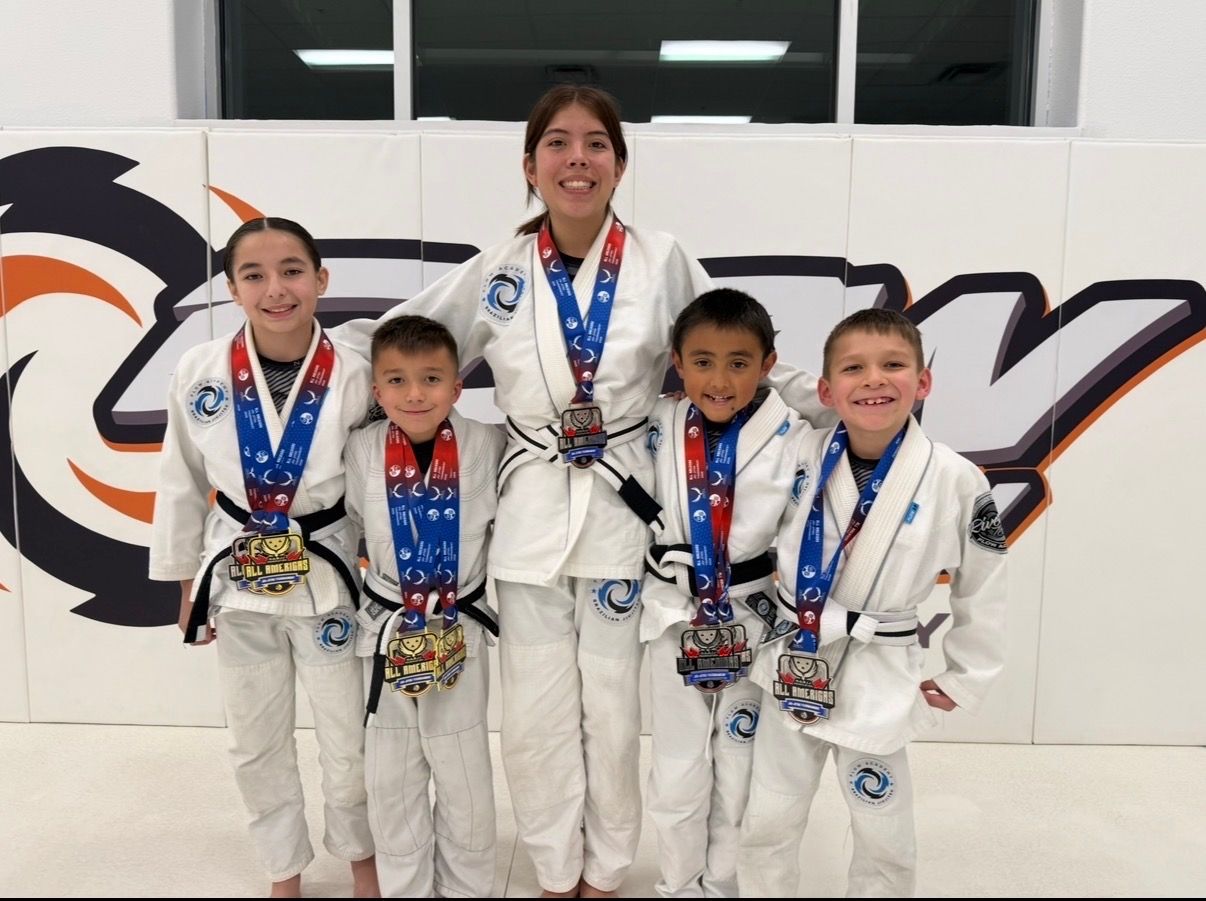 Five Flow Academy kids and youth in white gis smiling and wearing All Americas medals in front of the orange Flow wall logo