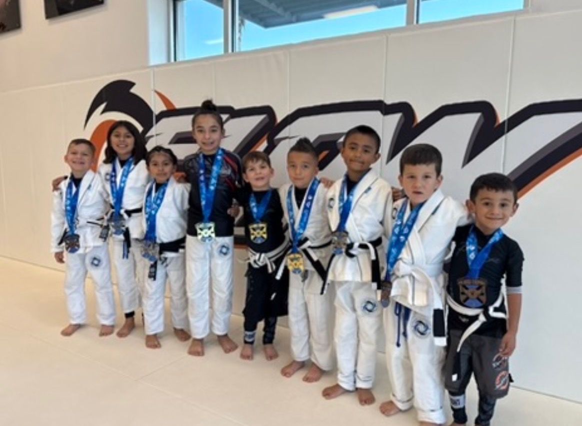 A row of young Flow Academy students in gis and rash guards wearing medals from a youth tournament, in front of the orange Flow wall logo