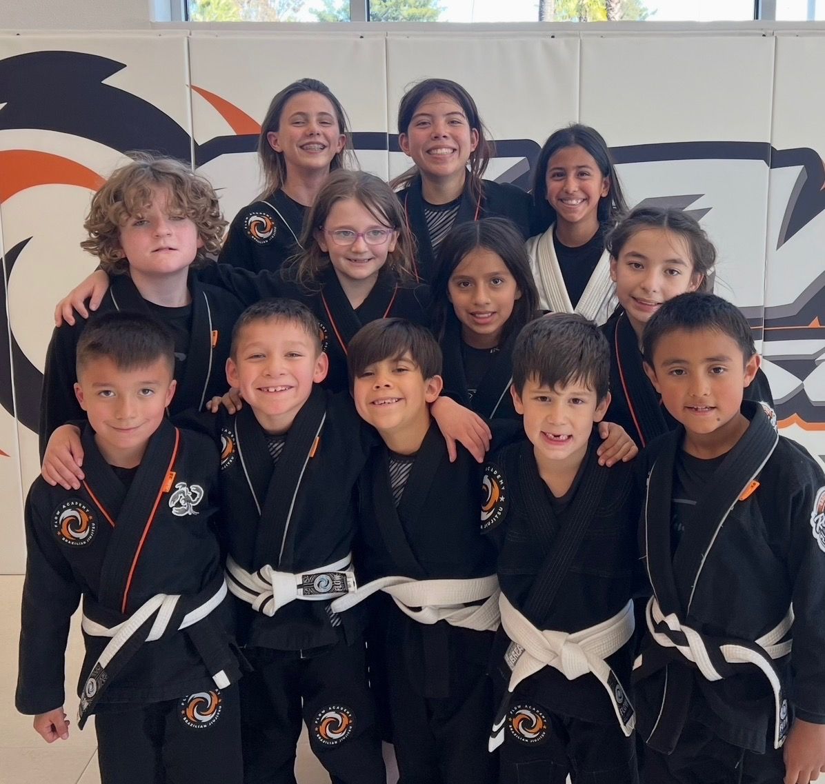 A group of smiling Flow Academy kids in black gis standing together in front of the orange Flow wall logo