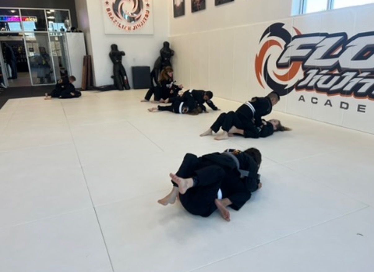 Students in black gis training live rounds in pairs across the mat, in front of the orange Flow wall logo