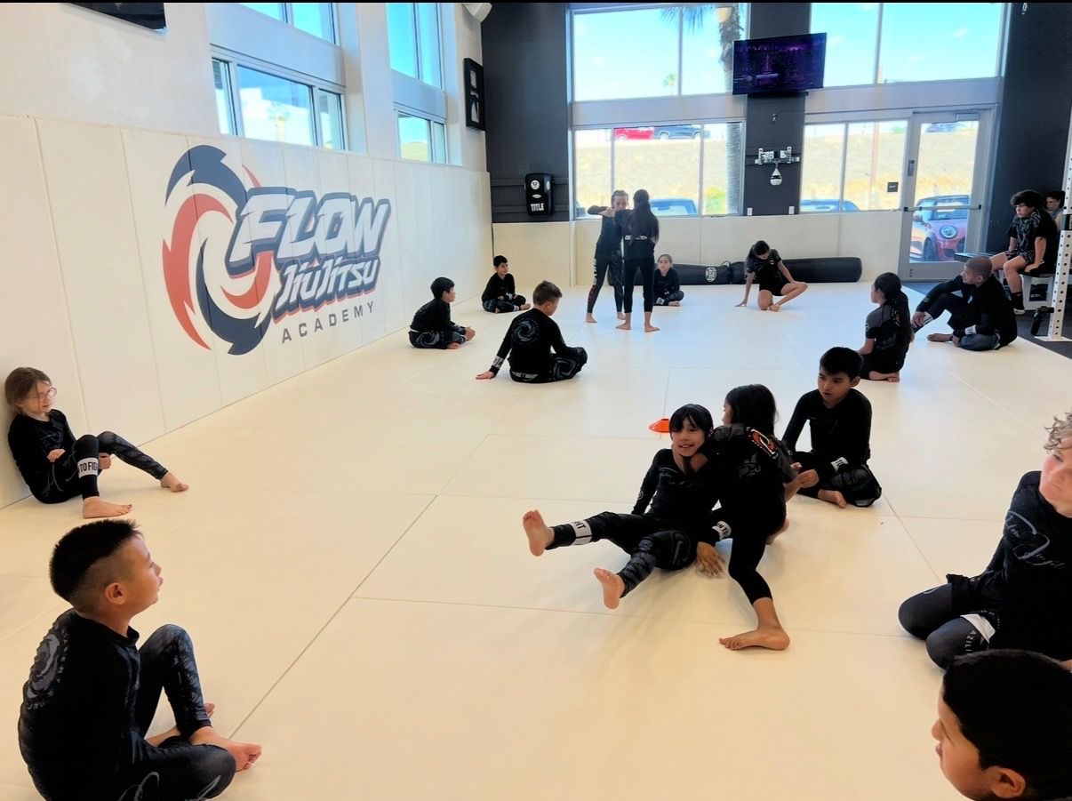 A kids class on the mat at Flow Academy, with students in rash guards seated around the room and two partners working together in the center