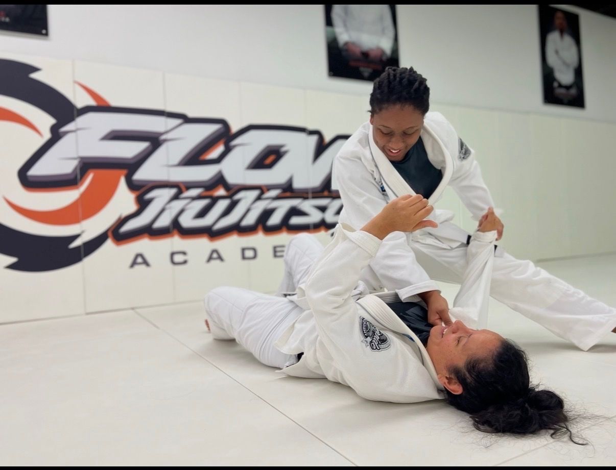 Two women in white gis practicing a jiu-jitsu escape on the mat in front of the orange Flow wall logo, both smiling