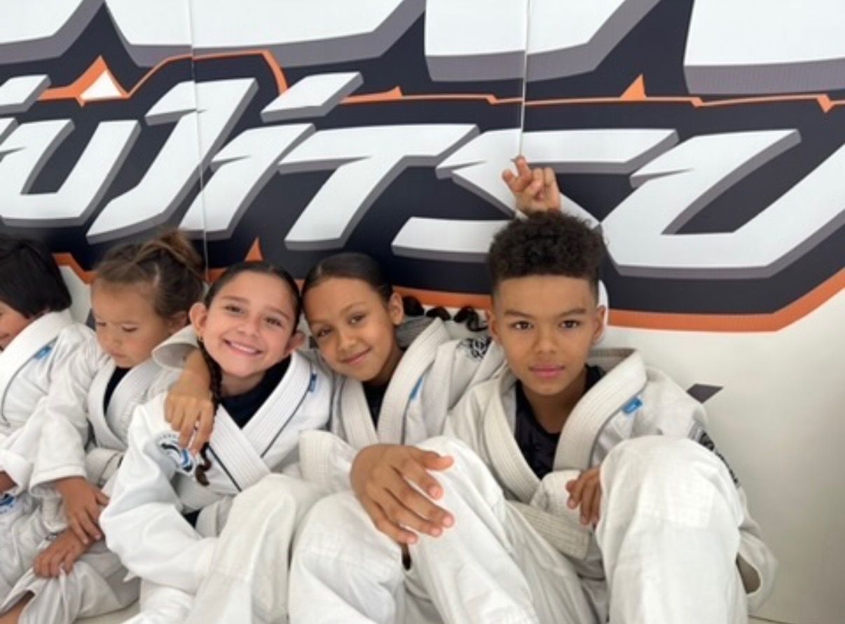 Flow Academy youth students in white gis sitting arm in arm and smiling against the orange Flow wall logo